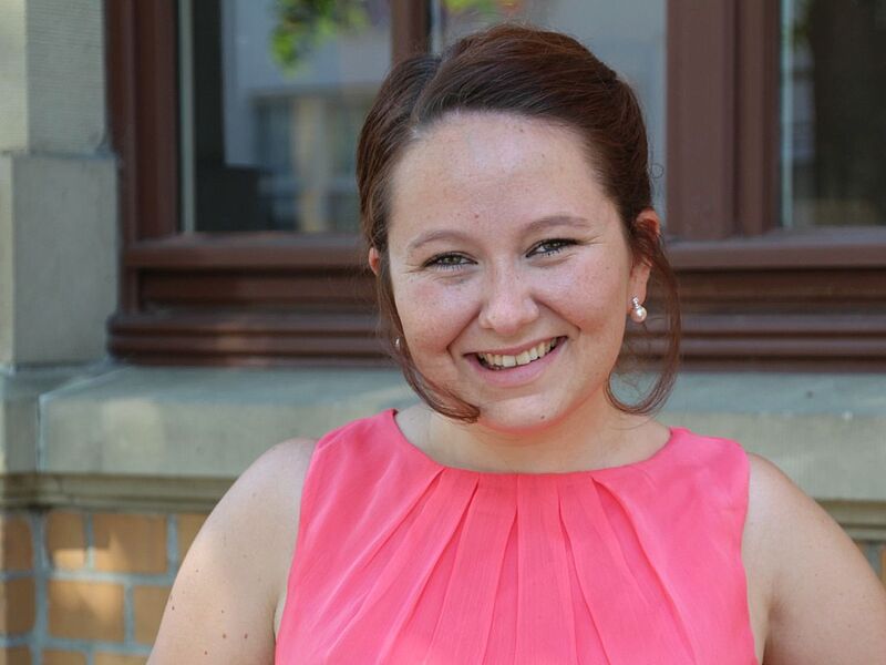 Eine lächelnde Frau mit hochgestecktem braunem Haar steht vor einem Gebäude mit Backsteinfassade und großen Fenstern. Sie trägt ein ärmelloses, pinkfarbenes Kleid mit Falten am Ausschnitt und Perlenohrringe.