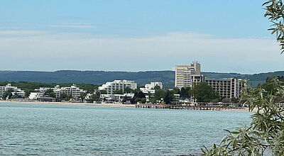 Blick über ein ruhiges Meer auf die bulgarische Küstenstadt Albena mit mehreren modernen Hotelgebäuden und einem langen Sandstrand, im Hintergrund bewaldete Hügel; im Vordergrund sind Zweige eines Baums sichtbar.