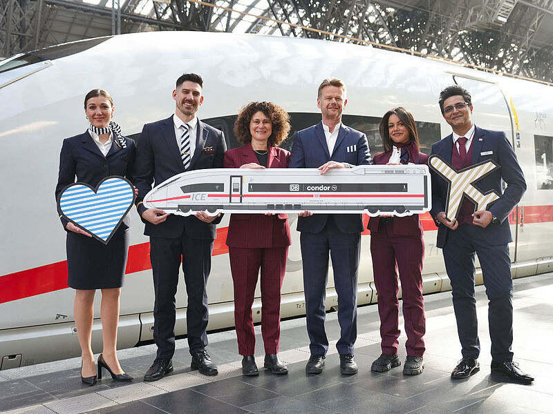 Sechs Airline- und Bahnmitarbeiter in Business-Kleidung posieren vor ICE mit Modellzug, Herz und Flugzeugsymbol, symbolisieren DB-Condor-Kooperation.