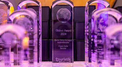  Mehrere transparente, lila beleuchtete Trophäen des „Globus Award 2024“ von touristik aktuell stehen aufgereiht auf einem Tisch. Auf der mittleren Auszeichnung ist die Gravur „Bester Sales Manager Außendienstler – Angela Distler, Iberostar“ zu erkennen.