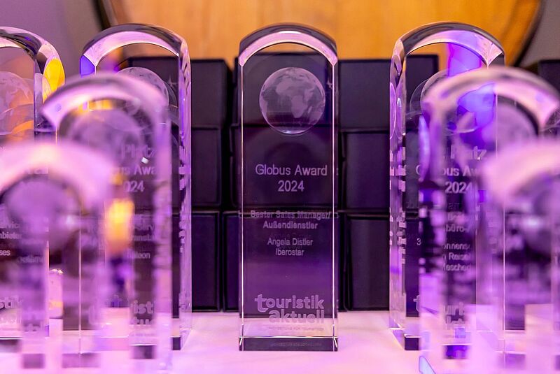  Mehrere transparente, lila beleuchtete Trophäen des „Globus Award 2024“ von touristik aktuell stehen aufgereiht auf einem Tisch. Auf der mittleren Auszeichnung ist die Gravur „Bester Sales Manager Außendienstler – Angela Distler, Iberostar“ zu erkennen.