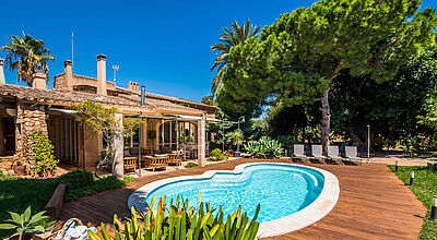 Ferienhaus-Urlaub, auf Mallorca auch gerne mit Pool, ist weiter im Kommen Ein mediterranes Ferienhaus auf Mallorca mit blauem Pool und Holzterrasse sowie einem Garten mit Palmen und Pinien bei sonnigem Sommerwetter mit blauem Himmel.