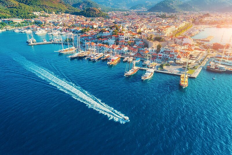 Luftaufnahme des Hafens von Marmaris, Türkei, bei Sonnenuntergang. Zahlreiche Segelboote und Yachten liegen an den Kais, während ein Schnellboot eine weiße Spur durch das tiefblaue Wasser zieht. Die Stadt erstreckt sich mit ihren weißen und roten Dächern entlang der Küste, eingerahmt von grünen Hügeln und Bergen. Historische Gebäude, darunter eine Burg, sind in der Altstadt zu sehen. Die warme Abendsonne taucht die Szenerie in ein goldenes Licht.