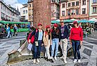 Eine Gruppe von fünf Frauen und einem Mann steht lächelnd vor einer Skulptur in einer belebten Altstadt mit historischen Gebäuden und Markständen. Alle tragen rote Kappen mit der Aufschrift „Emirates“, einige auch rote Kleidung oder Accessoires. Im Hintergrund sind Marktstände mit grün-weiß gestreiften Markisen und Passanten zu sehen. Die Stimmung ist freundlich und lebhaft bei Tageslicht.