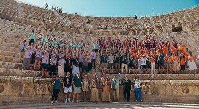 Eine große Gruppe von Menschen posiert auf den Stufen eines antiken Amphitheaters bei sonnigem Wetter. Die Teilnehmenden tragen bunte, farblich abgestimmte T-Shirts in Gruppenfarben (Lila, Türkis, Orange, Dunkelgrün) und heben jubelnd die Arme. Vorne stehen einige Personen in traditioneller Kleidung. Der Hintergrund zeigt die steinernen Sitzreihen des Theaters unter einem klaren blauen Himmel.