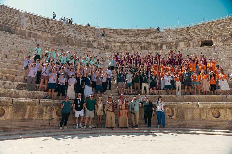 Eine große Gruppe von Menschen posiert auf den Stufen eines antiken Amphitheaters bei sonnigem Wetter. Die Teilnehmenden tragen bunte, farblich abgestimmte T-Shirts in Gruppenfarben (Lila, Türkis, Orange, Dunkelgrün) und heben jubelnd die Arme. Vorne stehen einige Personen in traditioneller Kleidung. Der Hintergrund zeigt die steinernen Sitzreihen des Theaters unter einem klaren blauen Himmel.