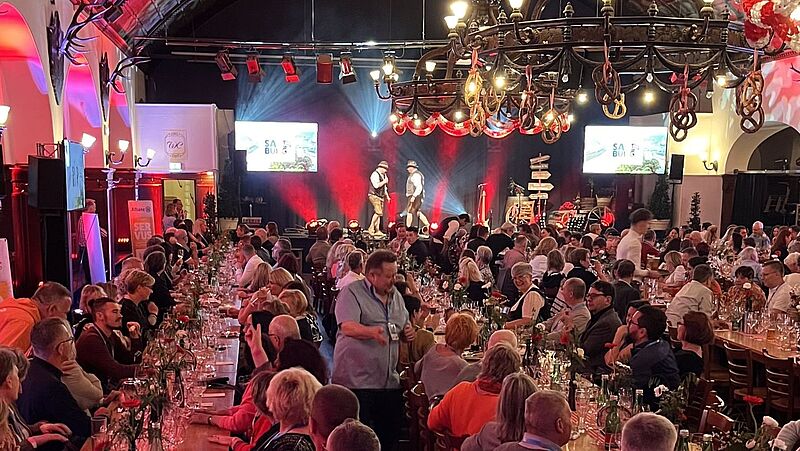 Ein festlich dekorierter Veranstaltungssaal mit langen, dicht besetzten Holztischen. Die Gäste genießen das Essen und unterhalten sich. Auf der Bühne stehen zwei Männer in traditioneller bayrischer Tracht und führen eine Darbietung auf. Der Saal ist mit roten Lichtern beleuchtet, an der Decke hängen Brezeln, und im Hintergrund sind Bildschirme mit der Aufschrift 'Salzburg' zu sehen