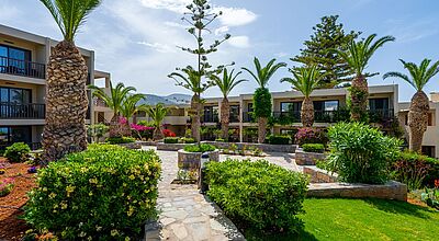Gepflegte Hotelanlage mit modernem, flachem Gebäude im mediterranen Stil, umgeben von Palmen, blühenden Sträuchern und gepflegten Steinwegen bei sonnigem Wetter auf Kreta