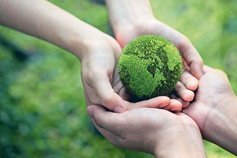 Mehrere Hände halten gemeinsam eine grüne, mit Gras bedeckte Weltkugel, die symbolisch die Erde darstellt. Der Hintergrund ist unscharf und zeigt eine natürliche, grüne Umgebung. Das Bild vermittelt ein Gefühl von Umweltschutz, Nachhaltigkeit und gemeinsamer Verantwortung für den Planeten.