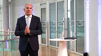 Lufthansa-Chef Carsten Spohr steht im Business-Anzug neben einem weißen Rednerpult in einem modernem Konferenzraum in der Lufthansa-Zentrale mit Glasfronten und blauer Beleuchtung.