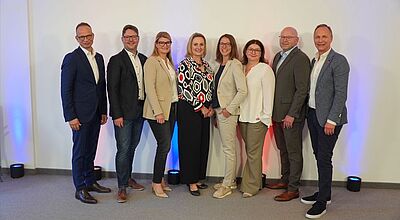 Acht Personen in Business-Kleidung stehen nebeneinander vor weißer Wand mit blau-roter LED-Beleuchtung. Professionelle Gruppenaufnahme.