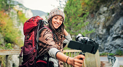 Eine junge Frau mit einem großen roten Wanderrucksack und einer bunten Strickmütze macht ein Selfie mit einer Kamera auf einem Stativgriff. Sie steht auf einer Brücke in einer malerischen Berglandschaft mit grünen Bäumen und Felsen im Hintergrund und lächelt fröhlich in die Kamera.