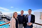 Eine Frau und zwei Männer lehnen sich an ein Geländer und lächeln in die Kamera. Im Hintergrund ist der Swimmingpool eines Kreuzfahrtschiffes zu sehen.