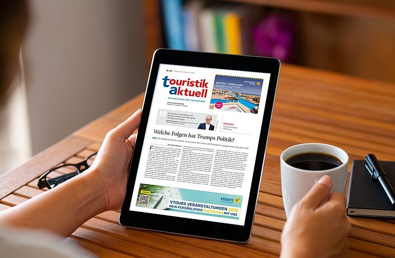   Eine Person hält ein Tablet mit einer digitalen Ausgabe der Fachzeitschrift 'touristik aktuell' in der Hand, während sie mit der anderen Hand eine Tasse mit schwarzen Kaffee hält. Die Zeitschrift auf dem Tablet enthält eine Schlagzeile über die Folgen von Trumps Politik. Auf einem Holztisch liegen zudem eine schwarze Brille, ein Notizbuch und ein Stift. Im Hintergrund sind verschwommene Bücher und ein violettes Objekt zu erkennen.   