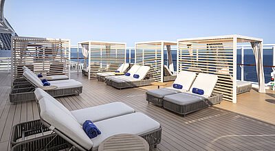 Das exklusive Sonnendeck des MSC Yacht Clubs, wie hier an Bord der MSC Seaview, findet sich künftig auch auf der Magnifica In einem exklusiven Außendeckbereich eines modernen Kreuzfahrtschiffes stehen vier bequeme, cremefarbene Doppel-Sonnenliegen. Auf ihren dicken Polsterauflagen sind jeweils zwei zusammengerollte blaue Handtücher drapiert.