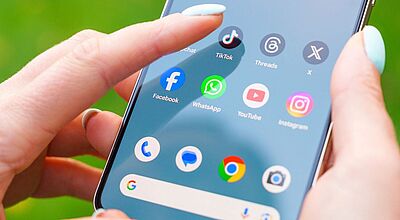 Nahaufnahme einer Hand mit hellblau lackierten Fingernägeln, die auf ein Smartphone-Display tippt, auf dem Social-Media-Apps wie TikTok, Facebook, WhatsApp, YouTube und Instagram zu sehen sind