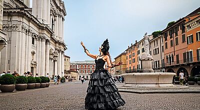 Eine Frau in einem eleganten schwarzen Ballkleid und einer schwarzen Feder-Maske steht auf einem historischen Platz mit Kopfsteinpflaster, hebt die Arme und blickt zum Himmel. Im Hintergrund sind eine barocke Kirche, bunte Altstadthäuser und eine Statue mit Brunnen zu sehen, während Passanten vorbeigehen.