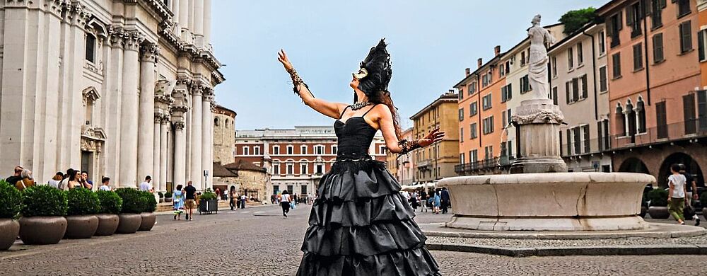 Eine Frau in einem eleganten schwarzen Ballkleid und einer schwarzen Feder-Maske steht auf einem historischen Platz mit Kopfsteinpflaster, hebt die Arme und blickt zum Himmel. Im Hintergrund sind eine barocke Kirche, bunte Altstadthäuser und eine Statue mit Brunnen zu sehen, während Passanten vorbeigehen.