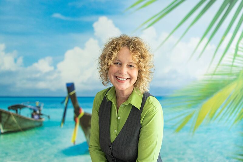Lächelnde Frau mit blonden Locken, hellgrüner Bluse und dunkelgrünem Pullunder, dahinter ein Bild mit zwei Booten im Meer