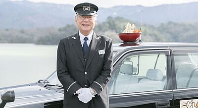 Lächelnder Chauffeur in schwarzer Uniform mit Mütze steht vor dunkler Limousine in Berglandschaft