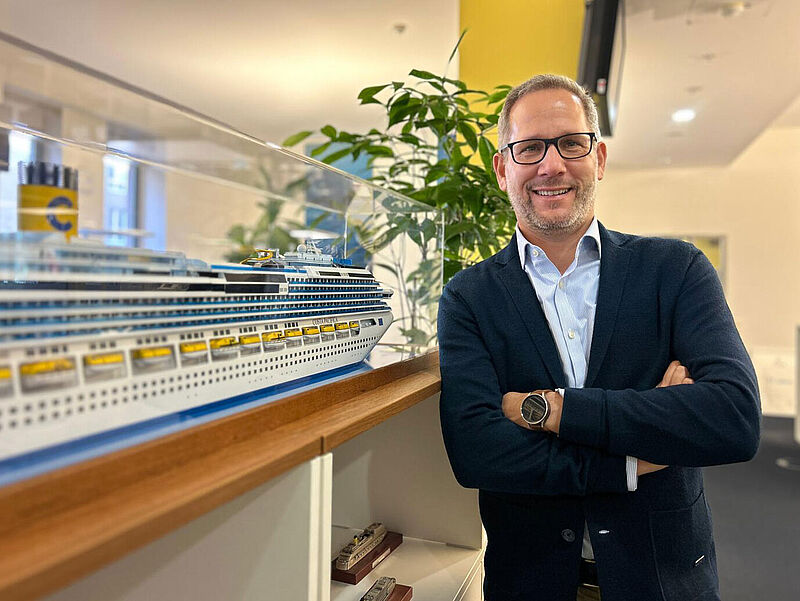Olaf Kistenmacher leitet seit gut einem Jahr den Vertrieb von Costa Kreuzfahrten. Foto: Costa Ein Reederei-Manager an die 50 posiert in der Unternehmenszentrale neben einem großen, in einer Glasvitrine stehenden Schiffsmodell und blickt freudig in die Kamera. Er hat kurze braune Haare, trägt eine Brille und ein dunkelblaues Sakko und hat die Arme verschränkt.