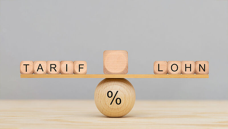 Von einer Tarifeinigung ist die Tourismusbranche noch weit entfernt Holzwürfel auf einer Wippe balancieren die Wörter „Tarif“ und „Lohn“, in der Mitte unter der Wippe befindet sich eine Holzkugel, auf der ein Prozentzeichen steht. Das Bild soll die Balance zwischen Tariflöhnen und tatsächlichen Gehältern symbolisieren.
