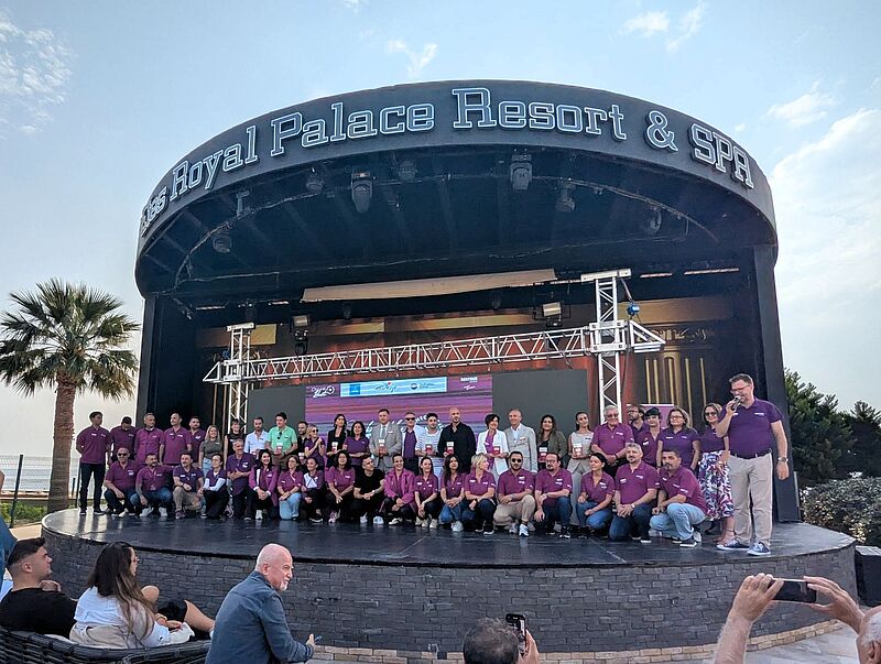 Auf der Bühne stehen zahlreiche Bentour-Mitarbeiter, in lila gekleidet, zusammen mit Reisebüro-Partnern, die einen Preis gewonnen haben, im Hintergrund blauer Himmel und Palmen.