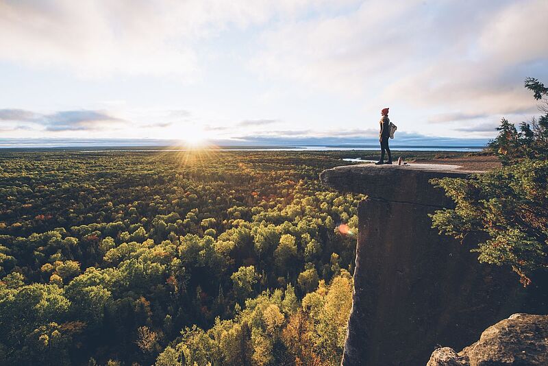 Junge Frau mit roter Pudelmütze blickt von einer Klippe über eine bewaldete herbstliche Landschaft