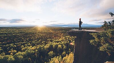 Junge Frau mit roter Pudelmütze blickt von einer Klippe über eine bewaldete herbstliche Landschaft