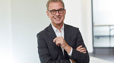 Mann mittleren Alters mit Brille und grauen Haaren, trägt einen dunklen Anzug und weißes Hemd, steht lächelnd mit verschränkten Armen in einem modernen Bürogebäude.