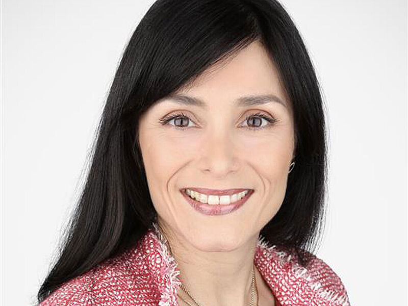 Porträt einer lächelnden Frau mittleren Alters mit langen schwarzen Haaren und dunklen Augen. Sie trägt einen rot-weiß-karierten Baumwollpullover. 