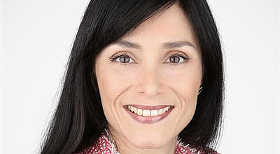 Porträt einer lächelnden Frau mittleren Alters mit langen schwarzen Haaren und dunklen Augen. Sie trägt einen rot-weiß-karierten Baumwollpullover. 