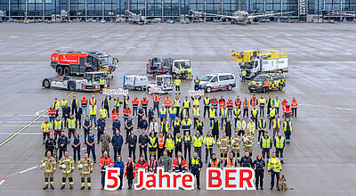 Beschäftigte des Flughafens Berlin stehen in einer Gruppe auf dem Vorfeld vor dem Terminalgebäude, vor sich halten sie Aufsteller, auf denen „5 Jahre BER“ steht.