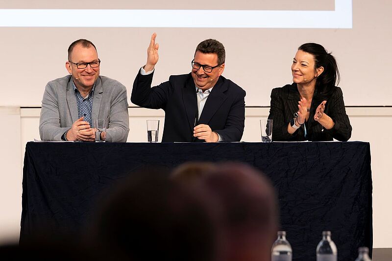 Ein Podium mit drei Personen, zwei Männer und eine Frau, die an einer Diskussion teilnehmen. Der Mann in der Mitte trägt einen dunklen Anzug, hebt eine Hand und lächelt, während die Frau rechts applaudiert und der Mann links lächelnd zuhört. Alle drei haben Mikrofone am Ohr. Das Publikum ist unscharf im Vordergrund zu sehen.