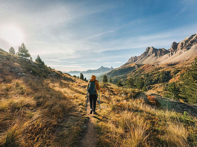 Frau in Rückansicht mit orangefarbener Jacke und Mütze sowie Rucksack und Trekking-Stöcken läuft durch eine Hochgebirgslandschaft