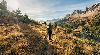 Noch bis zum 5. Dezember können Reisebüros das Verkaufsföderungspaket des Trekking-Spezialisten Hauser bestellen. Foto: Mumemories/istock Frau in Rückansicht mit orangefarbener Jacke und Mütze sowie Rucksack und Trekking-Stöcken läuft durch eine Hochgebirgslandschaft