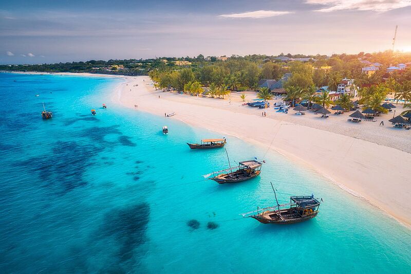Luftaufnahme der Fischerboote an der tropischen Meeresküste mit weißem Sandstrand bei Sonnenuntergang. Sommerurlaub am Indischen Ozean, Sansibar. Landschaft mit Boot, Palmen, transparentes blaues Wasser.