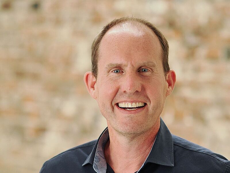 Am 1. Juni übernimmt Michael Geidel bei Gebeco die Verantwortung für den Vertrieb. Foto: Gebeco Porträt eines lächelnden Mannes mittleren Alters mit kurzen, leicht zurückweichenden braunen Haaren. Er trägt ein dunkles Hemd mit offenem Kragen. Der Hintergrund ist eine unscharfe, beige gemauerte Wand mit einer hellen Holzsäule an der rechten Seite des Bildes.