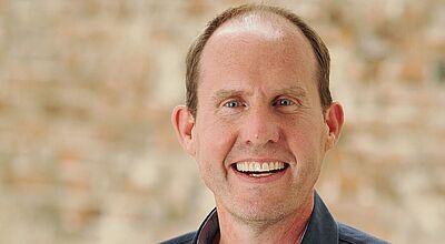 Porträt eines lächelnden Mannes mittleren Alters mit kurzen, leicht zurückweichenden braunen Haaren. Er trägt ein dunkles Hemd mit offenem Kragen. Der Hintergrund ist eine unscharfe, beige gemauerte Wand mit einer hellen Holzsäule an der rechten Seite des Bildes.