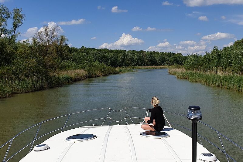 Eine blonde Frau, die nur mit dem Rücken zu sehen ist, sitzt auf dem Bug eines Bootes, das einen Fluss entlangfährt. Das Ufer ist geprägt von einer Naturlandschaft.