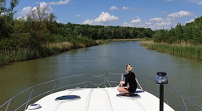 Foto: So sieht es aus, wenn Reisende auf dem Hausboot den Theiß entlangfahren. Foto: Riverly Eine blonde Frau, die nur mit dem Rücken zu sehen ist, sitzt auf dem Bug eines Bootes, das einen Fluss entlangfährt. Das Ufer ist geprägt von einer Naturlandschaft.