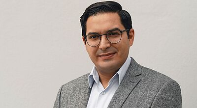 Porträt eines Mannes mit dunklem, seitlich gekämmtem Haar und runder Brille. Er trägt ein graues Sakko über einem hellen Hemd und blickt freundlich in die Kamera; neutraler, heller Hintergrund.