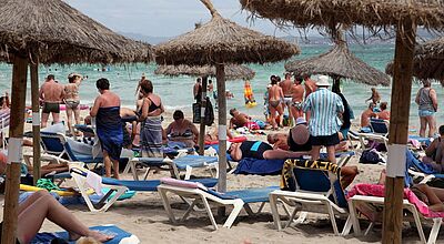 Bild von einem Strand mit ein paar Bastschirmen und dicht aneinandergereihten Liegestühlen auf dem viele Menschen in Badebekleidung zu sehen sind. Im Hintergrund ist das Meer zu erkennen.