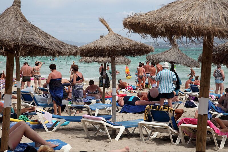Bild von einem Strand mit ein paar Bastschirmen und dicht aneinandergereihten Liegestühlen auf dem viele Menschen in Badebekleidung zu sehen sind. Im Hintergrund ist das Meer zu erkennen.