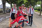 Vier fröhliche Menschen posieren im Freien für ein Foto, umgeben von Bäumen. Ein Mann trägt Pilotenuniformen mit weißen Hemden, Krawatten und Pilotenmützen. Eine Frau ist als Flugbegleiterin verkleidet und trägt ein rosa Hütchen, ein weißes Hemd und einen schwarzen Rock. Ein weiterer Mann steht im Vordergrund und trägt ein auffälliges, aufblasbares Flugzeugkostüm sowie ein rotes Trikot und eine rote Kappe. Ein weiterer Mann trägt eine bunte Kette aus künstlichen Blüten und einen Strohhut. Alle wirken gut gelaunt und scheinen sich in einer ausgelassenen, möglicherweise karnevalesken Stimmung zu befinden. Im Hintergrund sind weitere Menschen auf einer Bank sowie eine überdachte Holzhütte zu sehen.