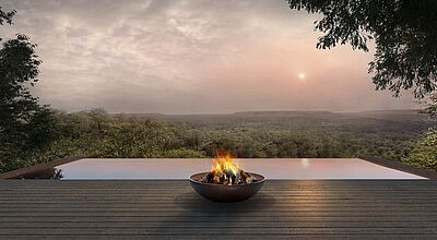 Panoramaansicht eines modernen, offenen Decks mit einem Feuerkorb im Vordergrund, der eine warme Atmosphäre erzeugt. Im Hintergrund erstreckt sich eine weitläufige, grüne Landschaft in den Dämmerungsstunden, während die untergehende Sonne den Himmel in sanften Farben erleuchtet. Perfektes Beispiel für luxuriöse, naturnahe Unterkünfte in Mosambik.