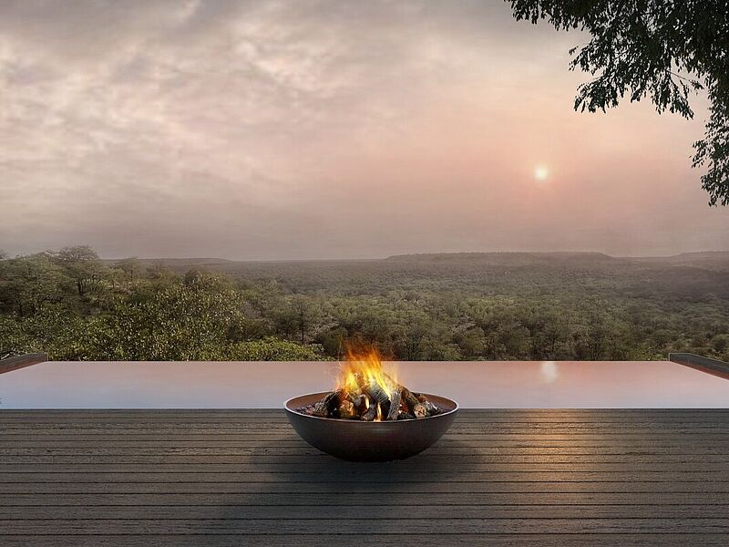 Panoramaansicht eines modernen, offenen Decks mit einem Feuerkorb im Vordergrund, der eine warme Atmosphäre erzeugt. Im Hintergrund erstreckt sich eine weitläufige, grüne Landschaft in den Dämmerungsstunden, während die untergehende Sonne den Himmel in sanften Farben erleuchtet. Perfektes Beispiel für luxuriöse, naturnahe Unterkünfte in Mosambik.