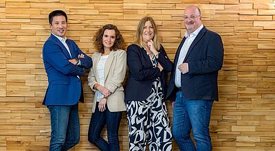 Vier Personen stehen lächelnd nebeneinander vor einer Wand aus hellen Holzpaneelen. Sie tragen Business-Casual-Kleidung – Blazer, Jeans und geschlossene Schuhe – und posieren selbstbewusst, teils mit verschränkten Armen. Der Boden ist aus dunklem Holz, und die Stimmung wirkt freundlich und professionell.