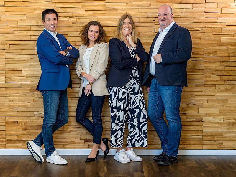 Vier Personen stehen lächelnd nebeneinander vor einer Wand aus hellen Holzpaneelen. Sie tragen Business-Casual-Kleidung – Blazer, Jeans und geschlossene Schuhe – und posieren selbstbewusst, teils mit verschränkten Armen. Der Boden ist aus dunklem Holz, und die Stimmung wirkt freundlich und professionell.