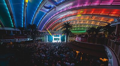 Blick auf ein Festival-Konzert im innenliegenden Beach Club eines großen Kreuzfahrtschiffes. Das Publikum steht im Dunkeln, das gebogene Hallendach ist bunt illuminiert, und weit hinten ist eine stark beleuchtete Bühne zu sehen.